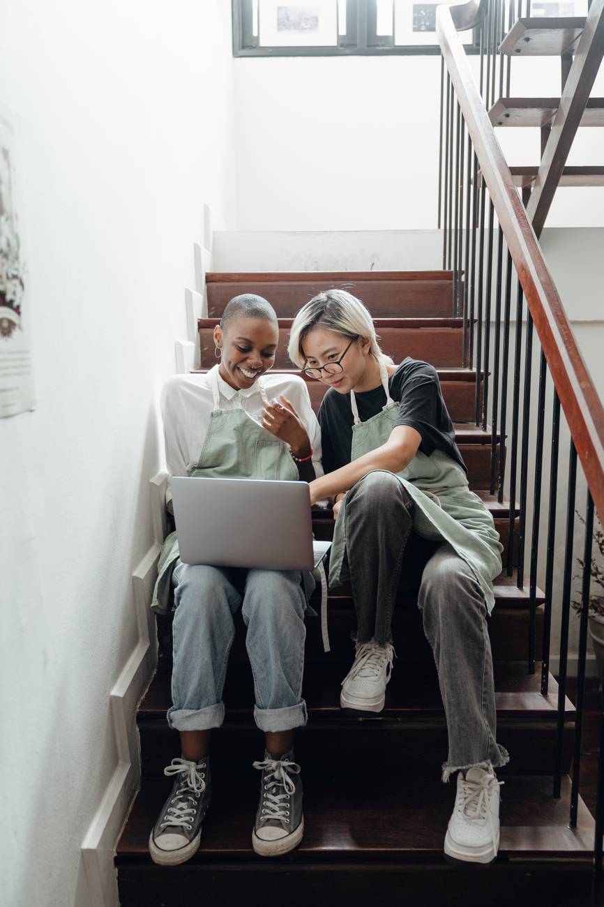 happy young multiethnic baristas watching movie on laptop sitting on stairs during break